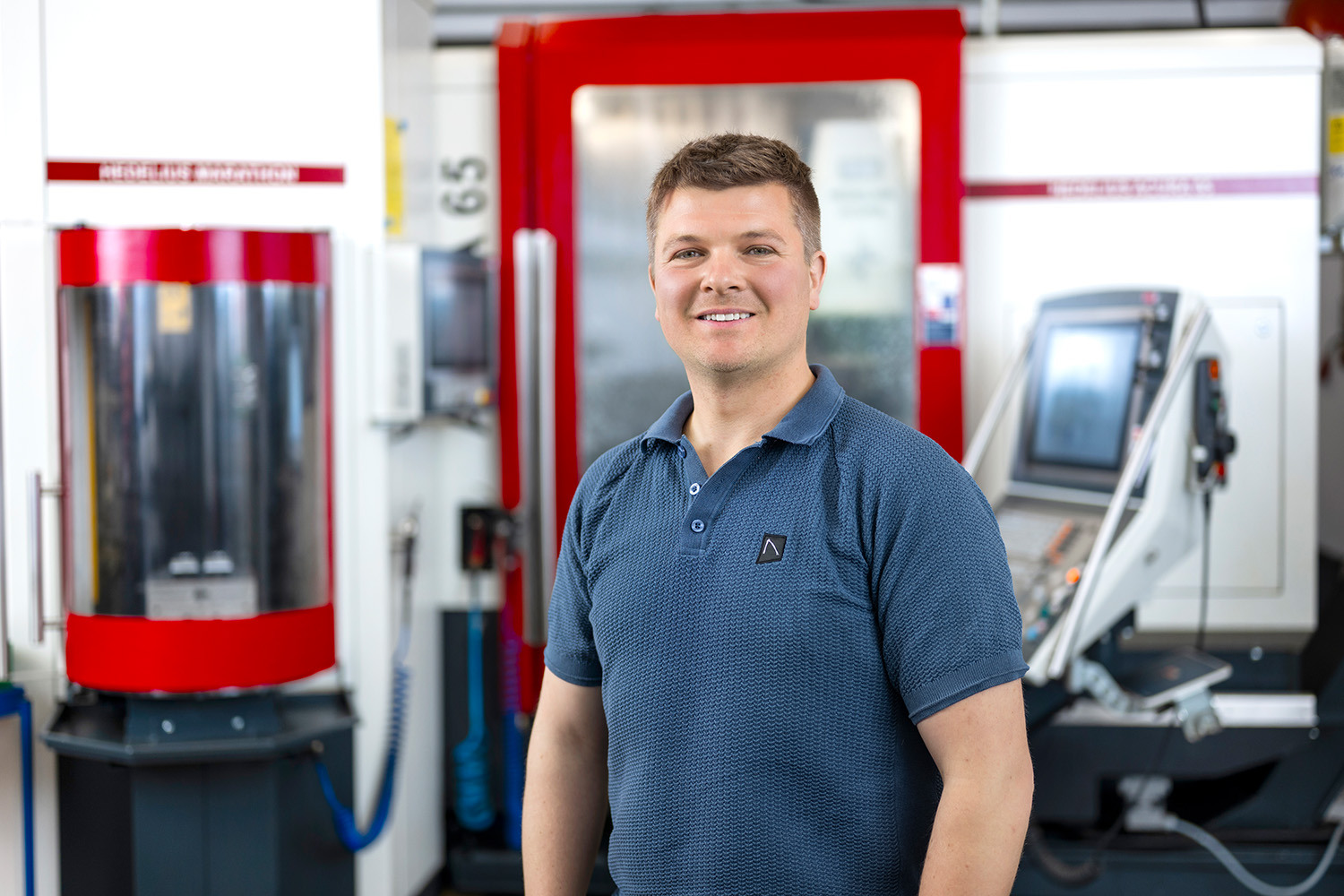 Ein Mann in einem blauen Polohemd steht lächelnd vor einer Maschine in einer industriellen Umgebung mit grauen und roten Elementen.