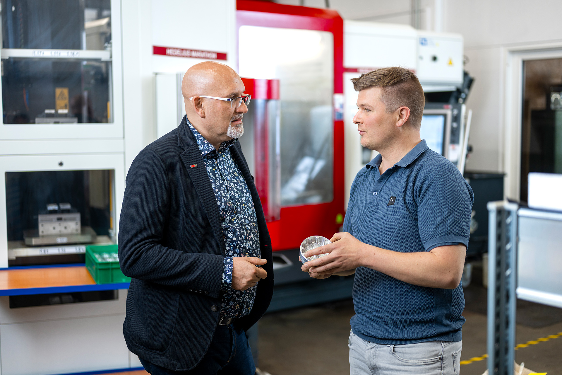 Zwei Männer in einem industriellen Umfeld, einer hält ein Objekt in der Hand, während sie sich unterhalten.