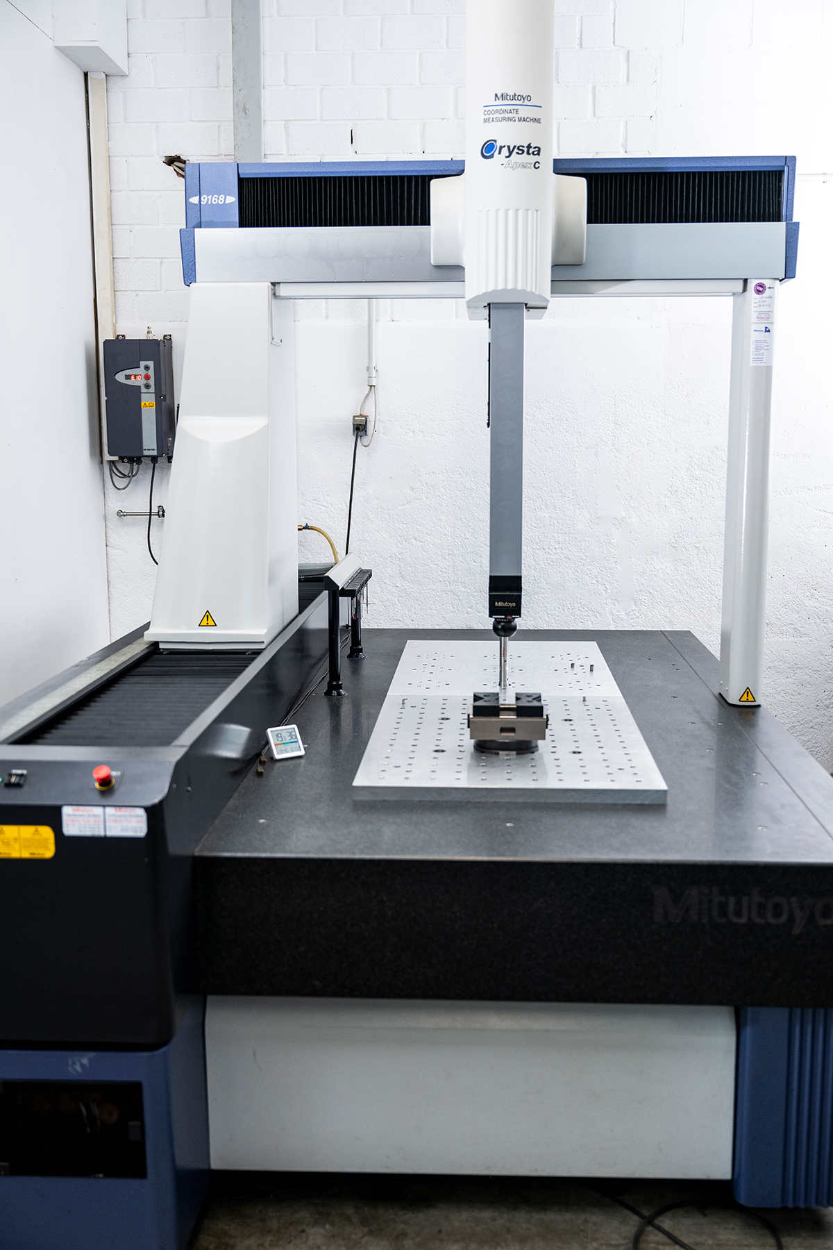 Eine Koordinatenmessmaschine der Marke Mitutoyo, Modell Crysta Apex C9168, in einem industriellen Raum mit weiß gestrichenen Wänden. Auf dem Messtisch befindet sich eine Aufspannvorrichtung für Werkstücke.